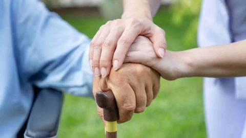 Nurse holding elderly man hand with cane on wheelchair in garden close up